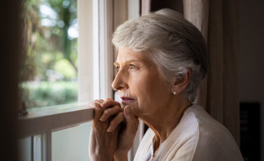A senior woman, who is experiencing signs of depression, looks out the window.
