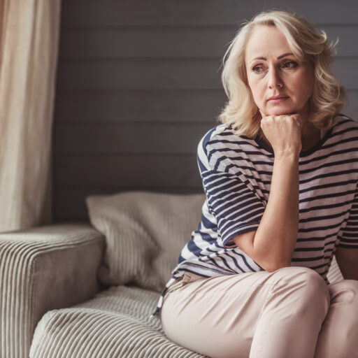 Stressed senior woman is leaning on her hand and looking downward while sitting on couch at home