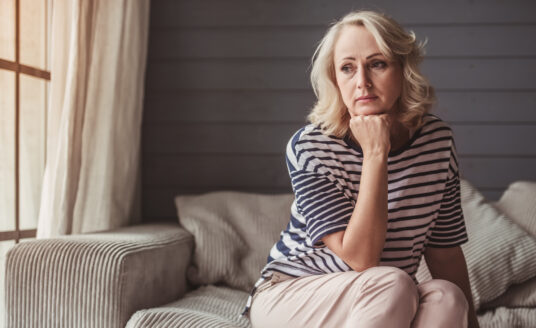 Stressed senior woman is leaning on her hand and looking downward while sitting on couch at home