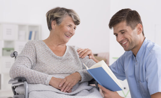 A volunteer at a skilled nursing home community reads to a senior woman.