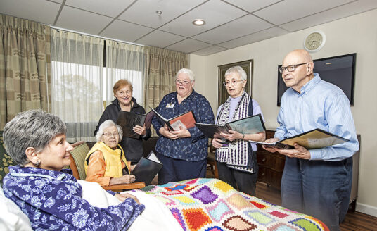 A group of volunteers sings to a senior woman because music touches lives of dying people.