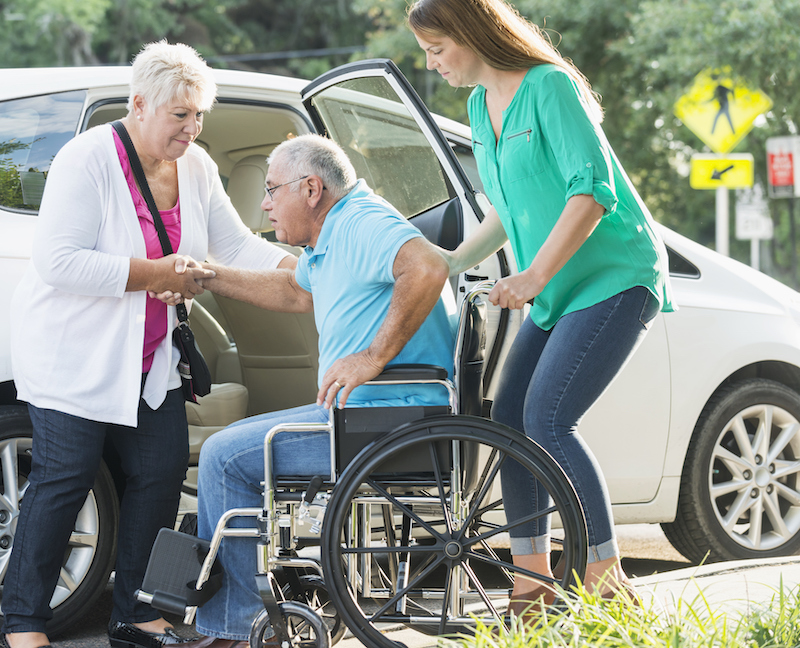 A wife and daughter help a senior man with differing care needs get into a car