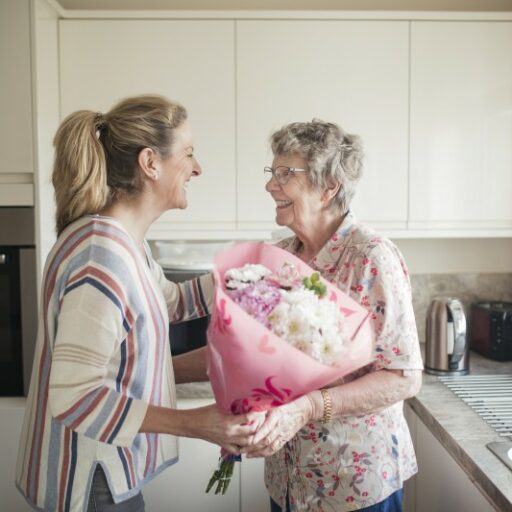 An older adult woman greets her senior mother, who is living with Alzheimer's, with a bouquet of flowers.