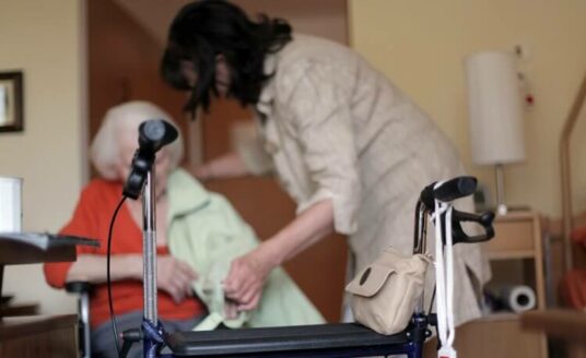 An adult woman helps her mother stand up with her walker.