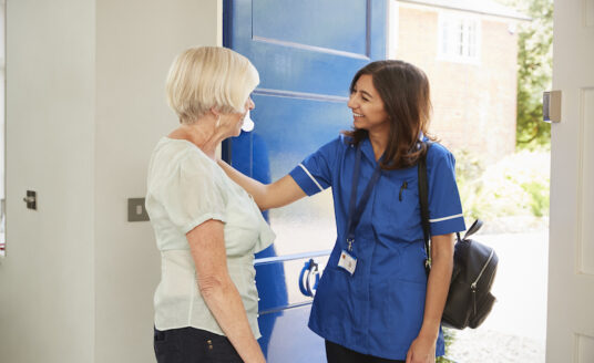A nurse greets a senior woman at her door, as she arrives to provide home care for the senior.