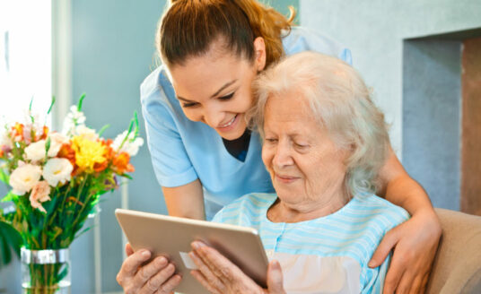 Respite care worker helps elderly woman use a tablet.