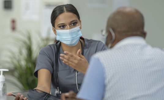 A senior patient and a female geriatric care manager meet in a medical clinic while wearing protective face masks to avoid the transfer of germs.