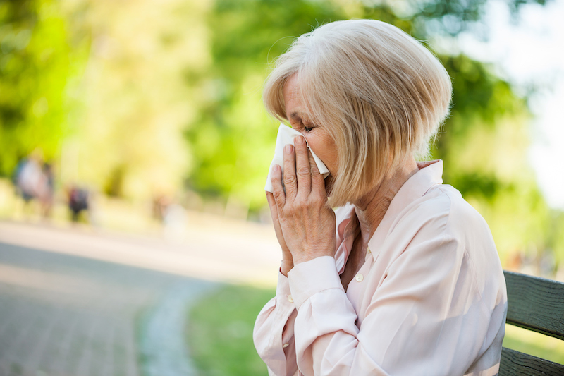 Adult woman is sitting in park and blowing nose. She is having an allergic reaction, a common spring health concern for seniors.