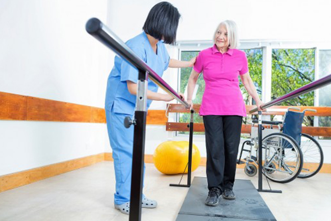 A senior woman in a bright pink shirt uses parallel bars to walk with assistance from a young female nurse