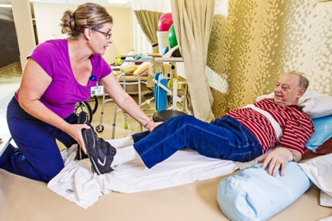 A middle aged white woman in a bright pink shirt helps an older white gentleman in a red striped shirt do physical therapy exercises with his legs.