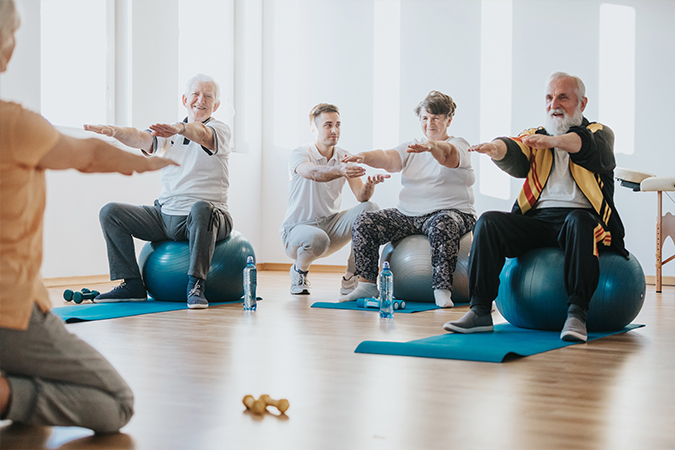 A group of three seniors sit on exercise balls with their arms out while a physical therapist attends to them