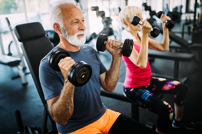 A senior man and woman work out on benches and lift dumbbells