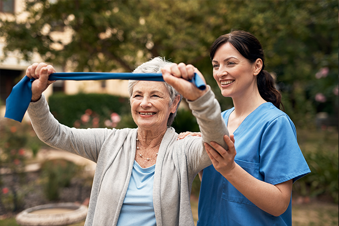 A young brunette woman in blue scrubs helps a senior woman exercise her arms with a resistance band