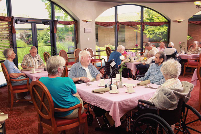 A group of seniors at Bethesda Southgate sit in the well-lit dining room, with beautiful windows and wooden floors