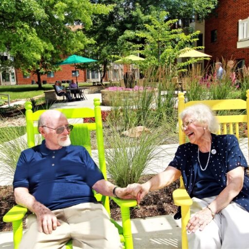A senior man and woman are seated in colorful chairs in the lush Bethesda Terrace courtyard | Bethesda Terrace allows seniors to receive care at home through Bethesda