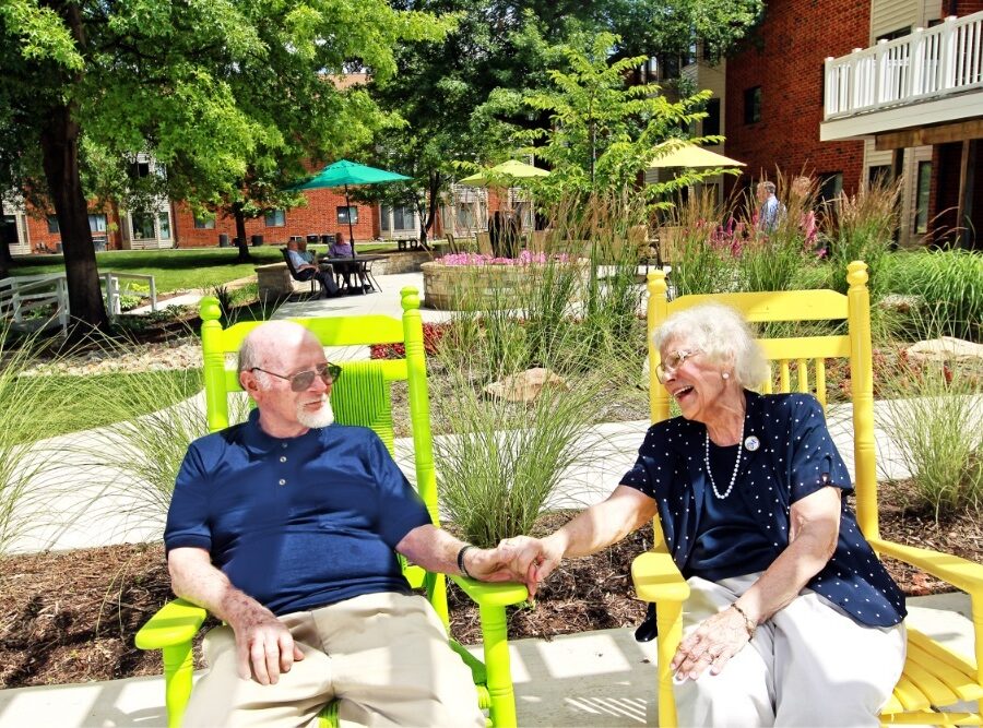 A senior man and woman are seated in colorful chairs in the lush Bethesda Terrace courtyard | Bethesda Terrace allows seniors to receive care at home through Bethesda