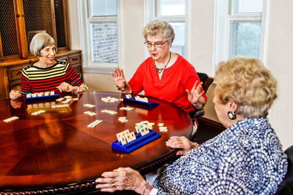 Three senior women play a game of Rummikub together at a round wooden table