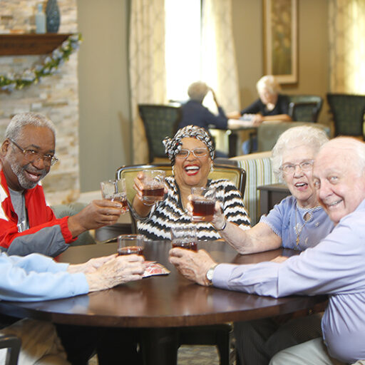 A group of five seniors raise their glasses toward the camera as they cheers in a comfortable living area around a round table