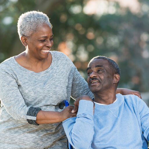 A black senior male in a wheelchair is accompanied by a similarly aged loved one who is female