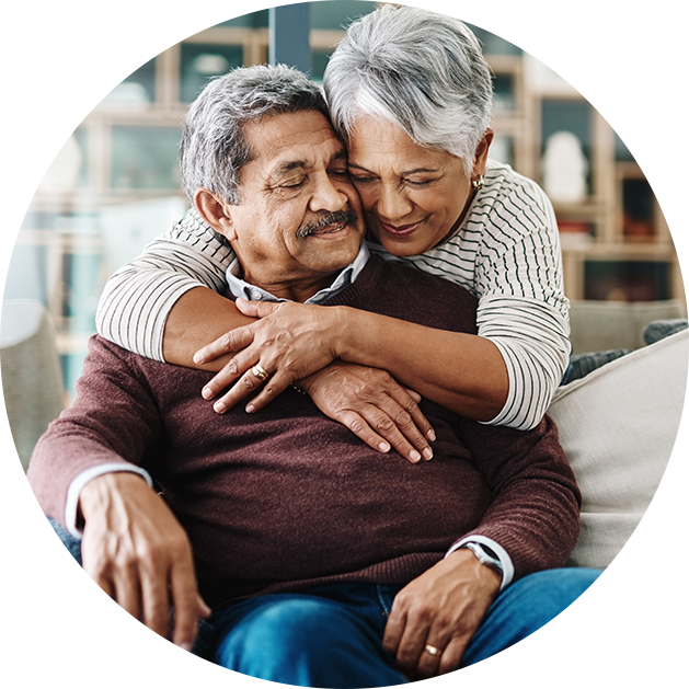 Senior woman hugs senior man from behind as he is seated on a couch