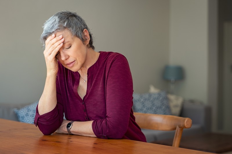 An older adult woman holds her head in her hand, a sign that she is stressed over her role as family caregiver, and may need to choose respite care.