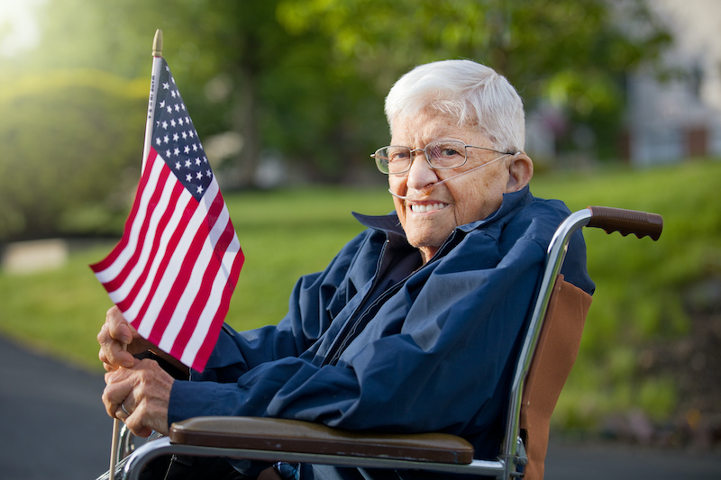 senior veteran sits in a wheelchair outside and holds an American flag