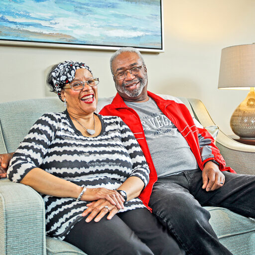 A senior man and woman pose and smile while seated on a loveseat