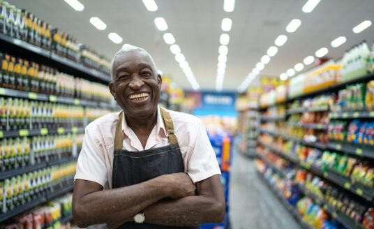 An older man stands in a grocery store aisle with an employee apron and arms crossed, representing the senior workforce