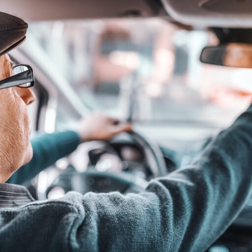 A male senior driver with a hat and glasses on adjusts his rear view mirror before driving.