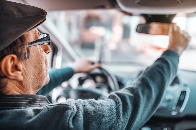 A male senior driver with a hat and glasses on adjusts his rear view mirror before driving.