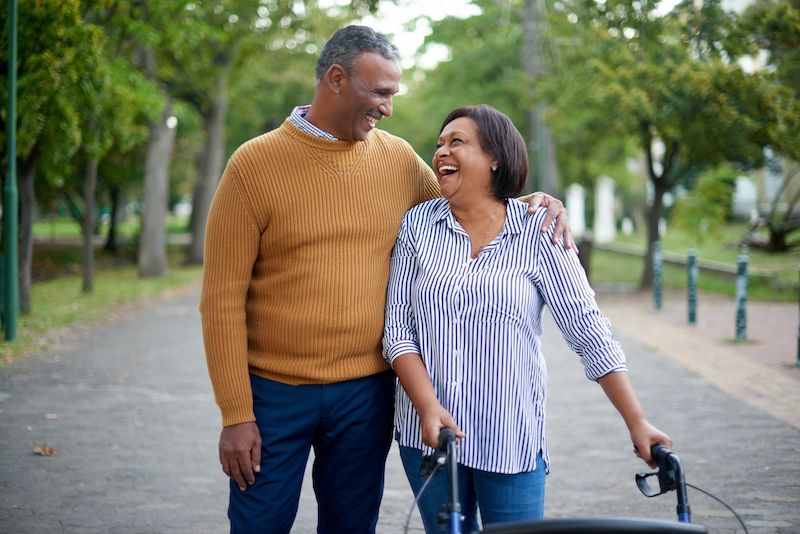 An older man and an older woman with a walker due to limited mobility take a stroll down a street lined with green trees