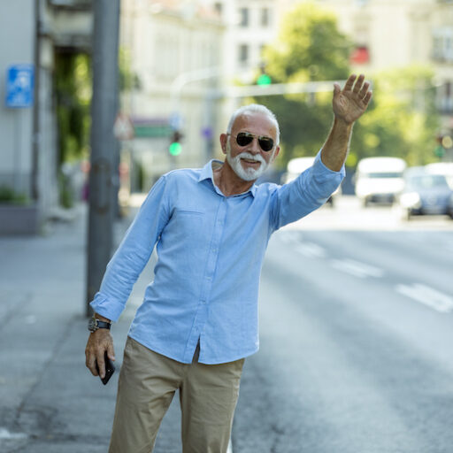 Senior man wearing sunglasses hails a ride on a sidewalk next to a busy street