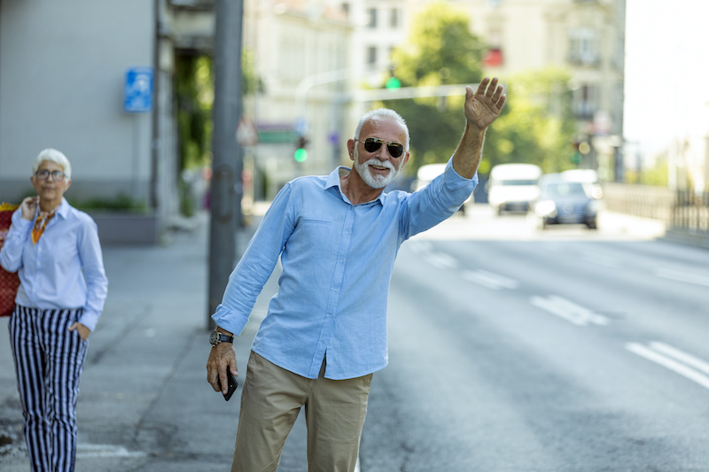 Senior man wearing sunglasses hails a ride on a sidewalk next to a busy street