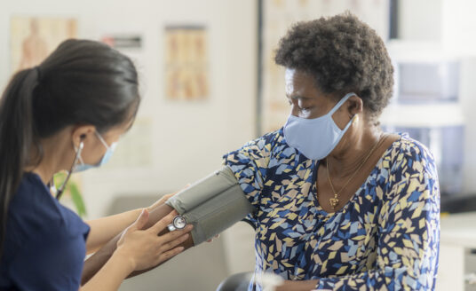 Senior woman wearing a face mask prevents hypertension by getting her blood pressure checked by a young female nurse