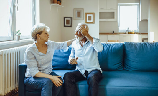 Senior woman and man sit on a couch, the woman tends to the man who is holding his head due to pain from a traumatic brain injury