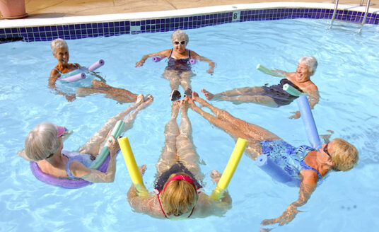 Six seniors working together in the pool as they perform a water aerobics routine, which is one of many great recreational sports for seniors