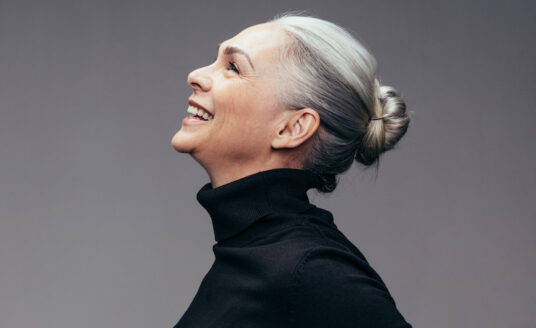 A senior woman in a black turtleneck smiles and tilt her head back in laughter, keeping a positive mindset.