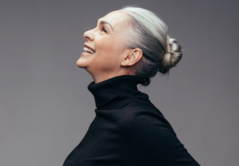 A senior woman in a black turtleneck smiles and tilt her head back in laughter, keeping a positive mindset.