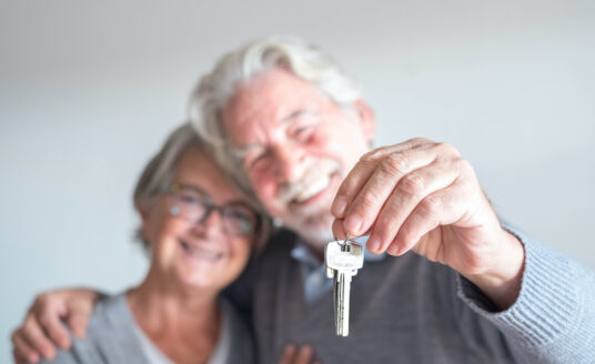 A senior man and woman, with the man's arm around the woman's shoulder. The man is holding a key, because the two of them are selling a home.