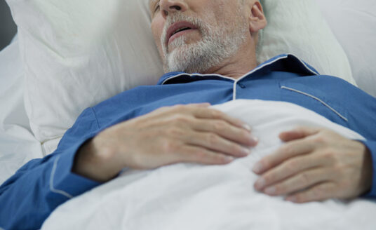 A senior man is tucked into bed, sleeping with his hands on his chest.