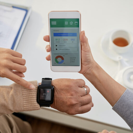 A senior woman receives assistance with a wearable health-monitoring device on her wrist, and looks at the corresponding apps for seniors on her phone