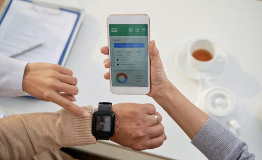 A senior woman receives assistance with a wearable health-monitoring device on her wrist, and looks at the corresponding app on her phone