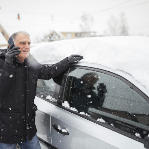 A senior man brushes snow off of his car before driving using winter driving tips