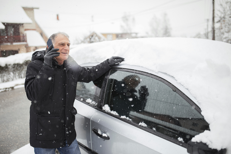A senior man brushes snow off of his car before driving using winter driving tips