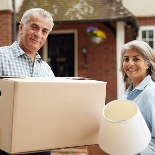 A senior man and woman are downsizing and moving to a new home, holding a box and a lamp in their hands.