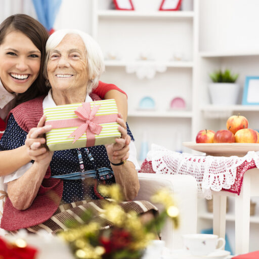An older woman is hugged from behind by a younger woman during the holidays, holding a wrapped gift and smiling despite holiday stress