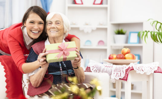 An older woman is hugged from behind by a younger woman during the holidays, holding a wrapped gift and smiling despite holiday stress