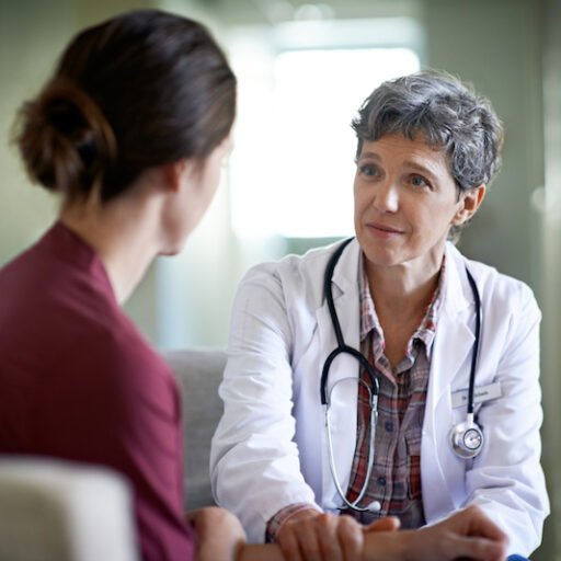 A woman has a conversation on a couch with an older female doctor, who is a member of the woman's care partner team.