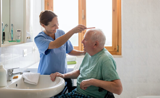 A senior man with Parkinsons disease gets help combing his hair from a young female caregiver.
