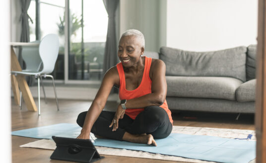 A senior woman practices self-care by preparing to do a yoga routine on a yoga mat in her living room.